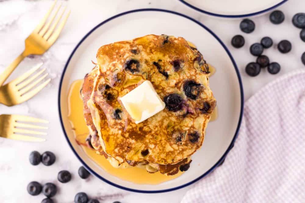 top view of a round white plate with a blue outer edge with a stack of fluffy blueberry pancakes with butter and syrup