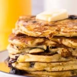 side view of a round white plate with a blue outer edge with a stack of fluffy blueberry pancakes with butter and syrup