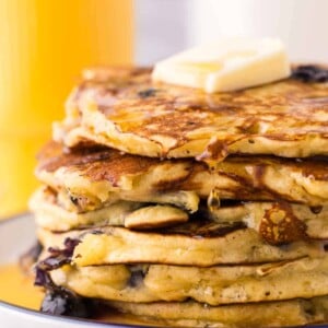 side view of a round white plate with a blue outer edge with a stack of fluffy blueberry pancakes with butter and syrup