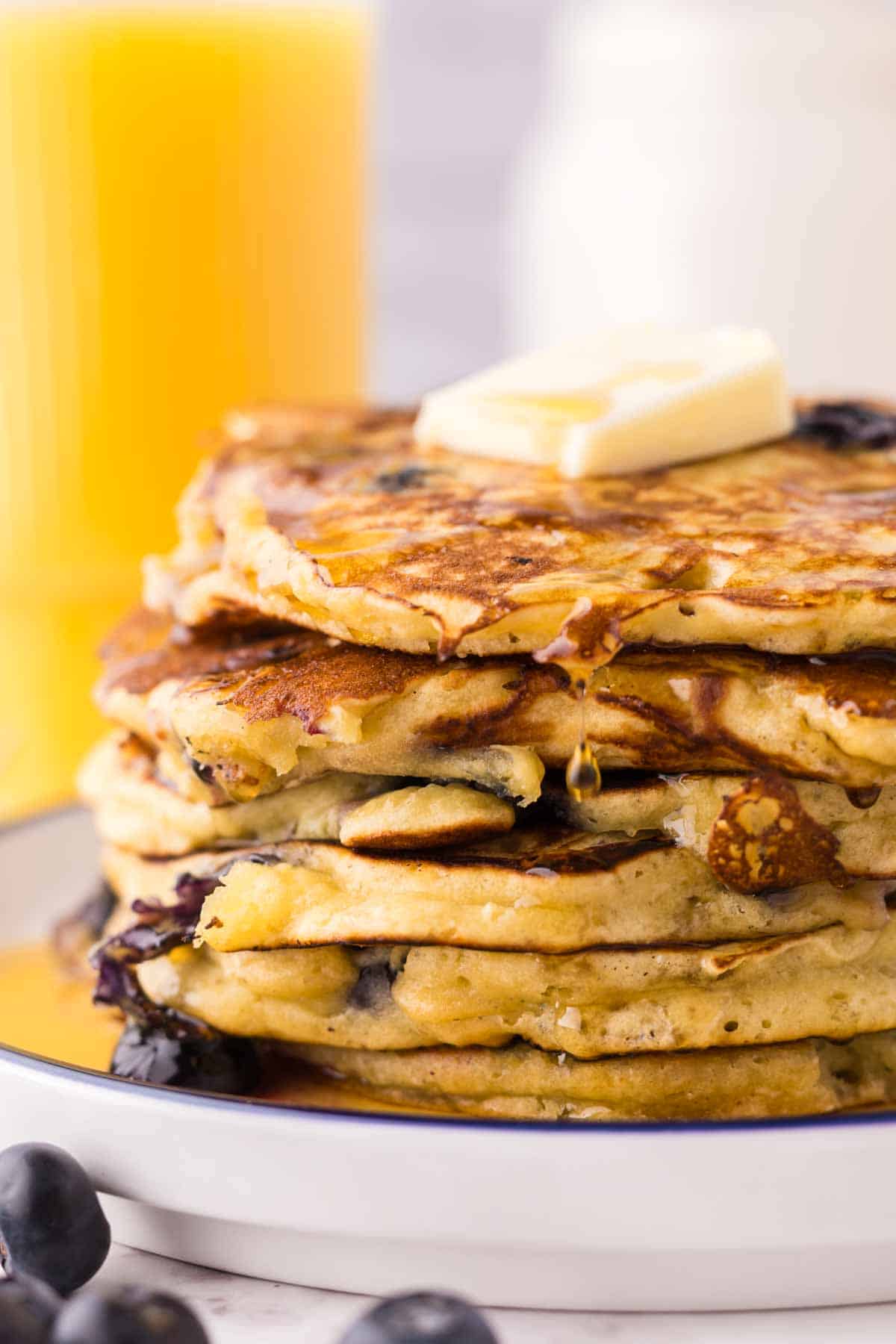 side view of a round white plate with a blue outer edge with a stack of fluffy blueberry pancakes with butter and syrup