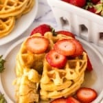 A stack of waffles from a buttermilk waffle recipe is topped with sliced strawberries and served on a white plate. A bite is cut out onto a fork, with more waffles and a basket of fresh strawberries waiting in the background.