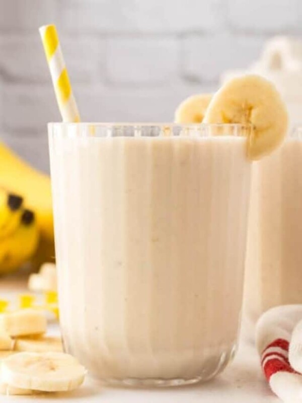 side view of a ribbed glass with a creamy banana smoothie with fresh bananas and a yellow striped paper straw two others in th backgrounders