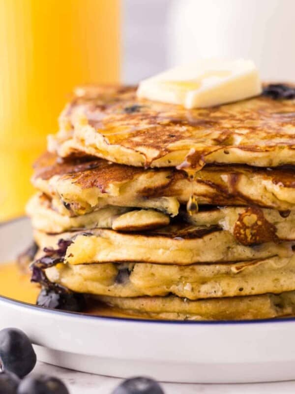 side view of a round white plate with a blue outer edge with a stack of fluffy blueberry pancakes with butter and syrup.