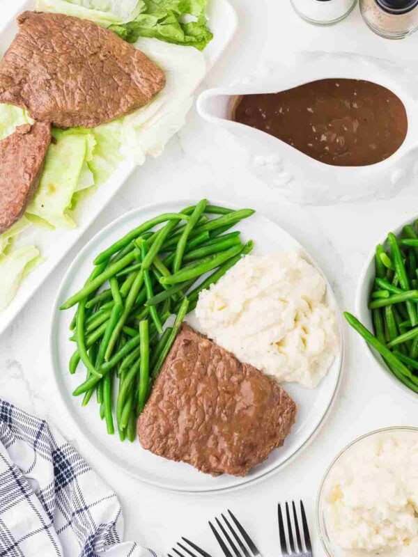 cooked round beef steak with green beens and mashed potatoes on a dinner plate with gravy to the side.
