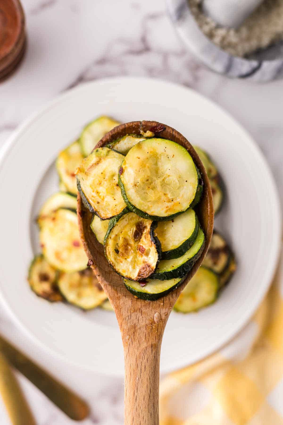 air fryer zucchini on a white plate being scooped up by a wooden spoon.