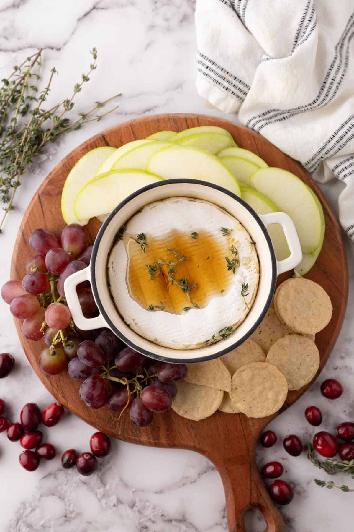baked brie wheel in a small round pot with thyme and honey next to crackers and fruit on a wooden board.