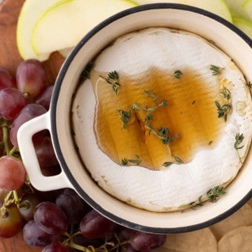 baked brie wheel in a small round pot with thyme and honey next to crackers and fruit on a wooden board.