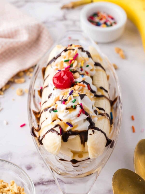 oval dish with classic banana split recipe with vanilla ice cream chocolate sprinkles and a cherry on top.