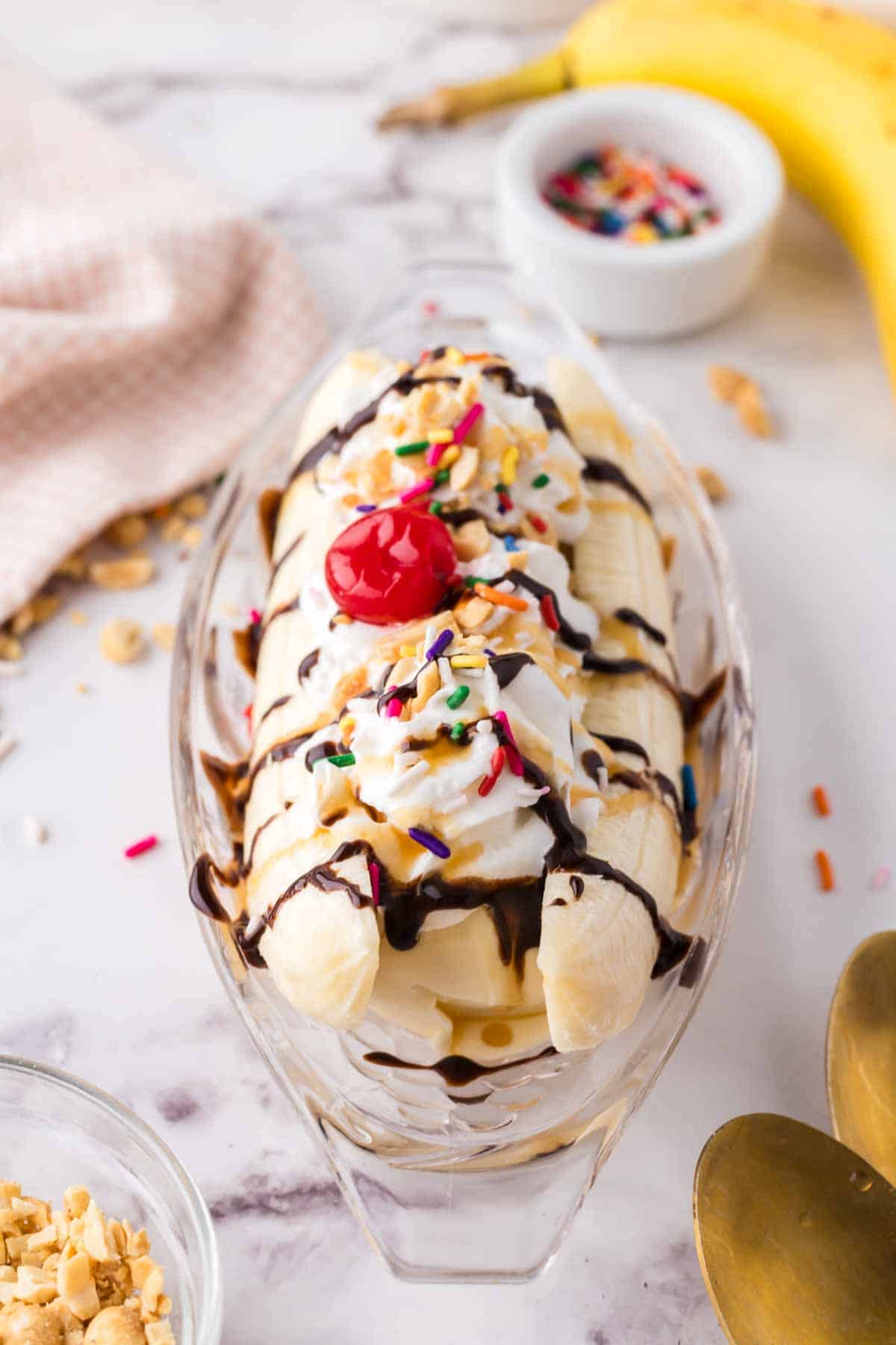 oval dish with classic banana split recipe with vanilla ice cream chocolate sprinkles and a cherry on top.