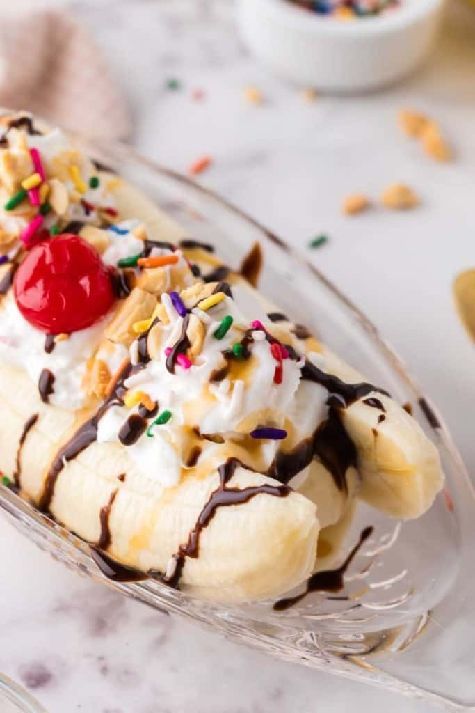 oval dish with classic banana split recipe with vanilla ice cream chocolate sprinkles and a cherry on top.