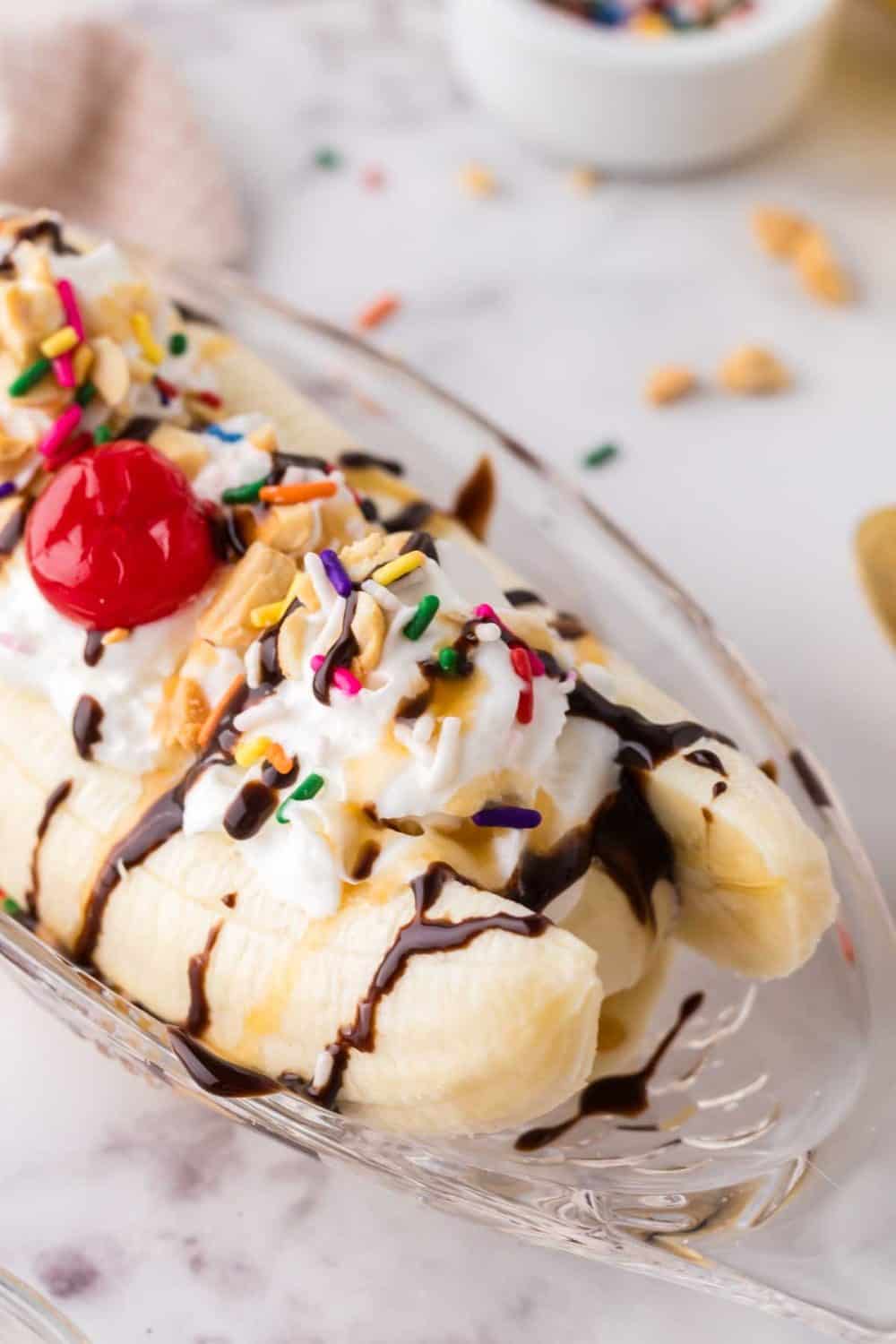 oval dish with classic banana split recipe with vanilla ice cream chocolate sprinkles and a cherry on top.