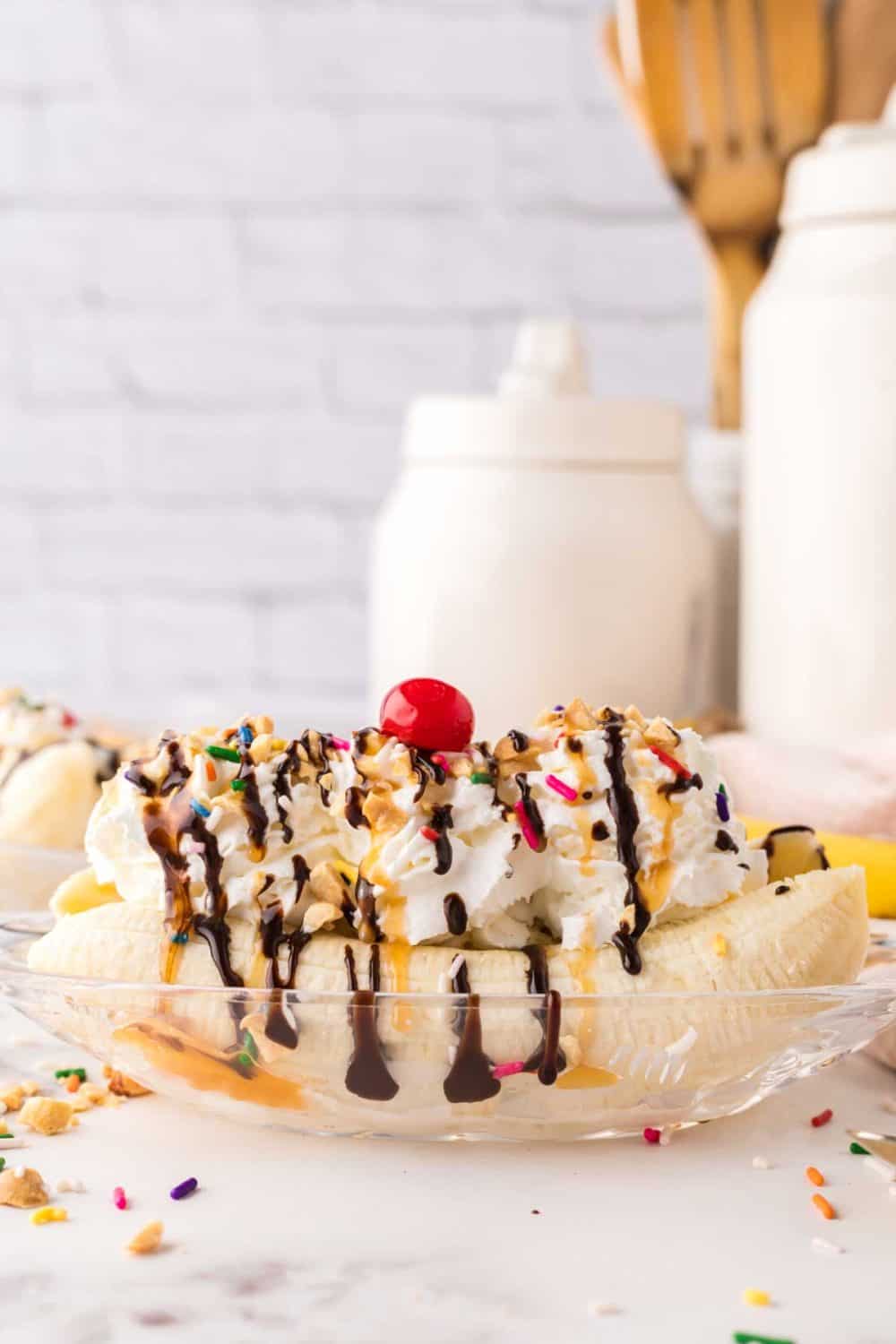oval dish with classic banana split recipe with vanilla ice cream chocolate sprinkles and a cherry on top.