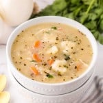 A bowl of creamy soup with vegetables and fluffy Bisquick dumplings, garnished with herbs, sits on a white napkin beside gold spoons, fresh parsley, and an onion on a marble surface.