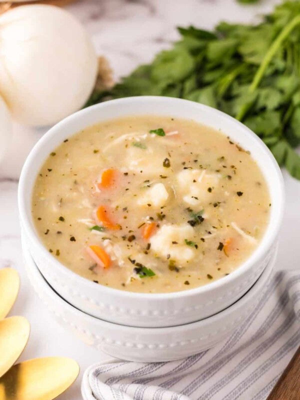 A bowl of creamy soup with vegetables and fluffy Bisquick dumplings, garnished with herbs, sits on a white napkin beside gold spoons, fresh parsley, and an onion on a marble surface.