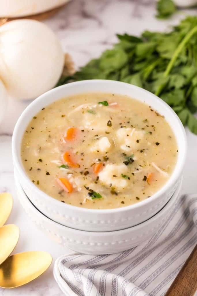 A bowl of creamy soup with vegetables and fluffy Bisquick dumplings, garnished with herbs, sits on a white napkin beside gold spoons, fresh parsley, and an onion on a marble surface.