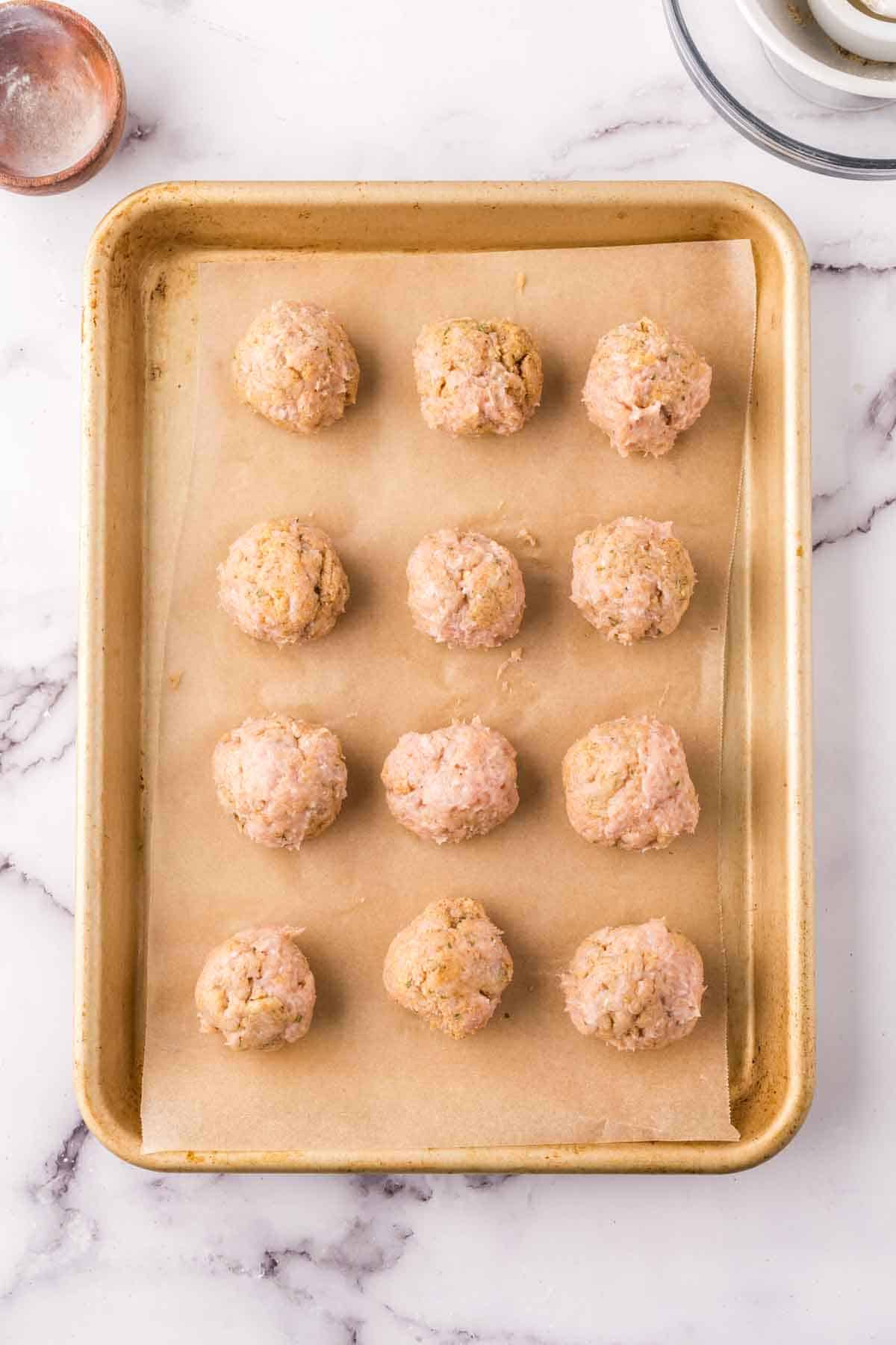 raw chicken meatballs lined on parchment on a baking tray.