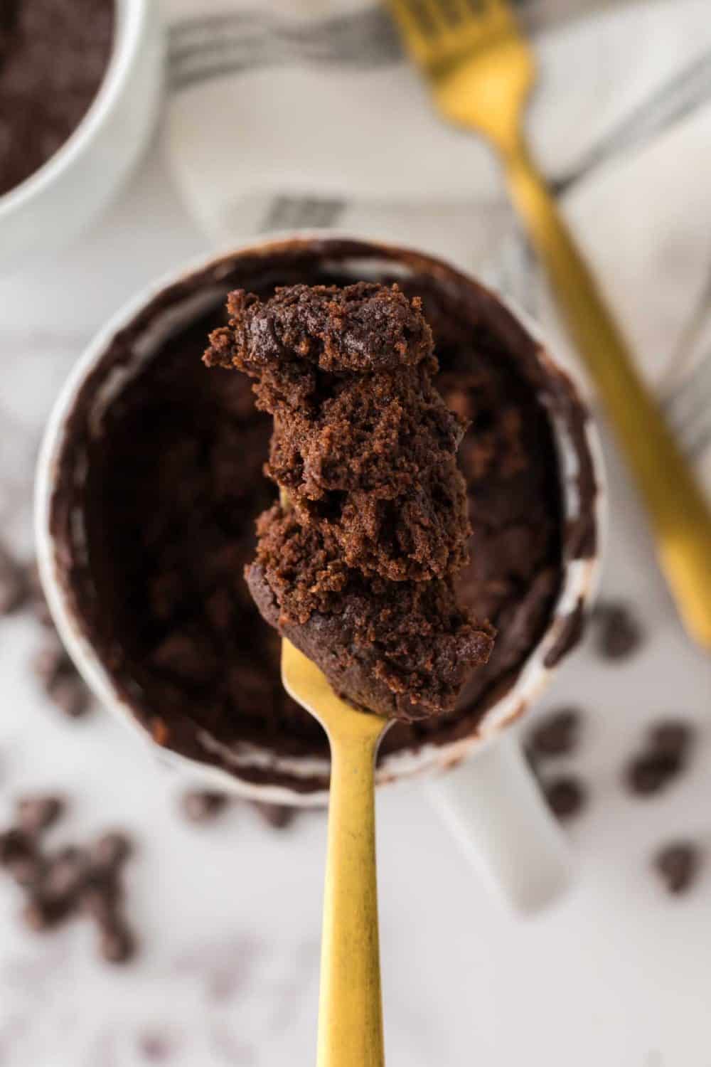 brown chocolate cake baked into a white coffee mug with chocolate chips around and a golden fork.