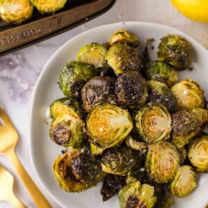 Oven Baked Brussels Sprouts on a round white plate next to whole lemons and golden forks.