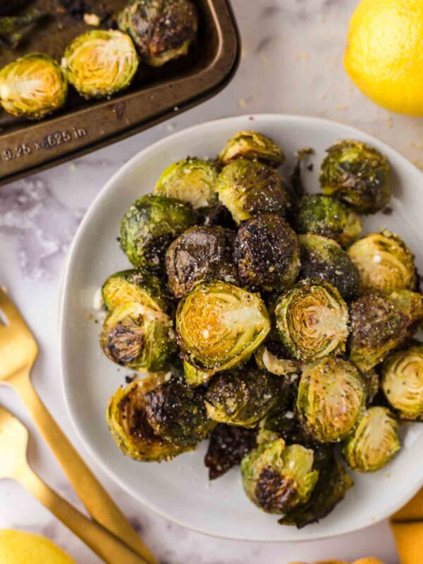 Oven Baked Brussels Sprouts on a round white plate next to whole lemons and golden forks.