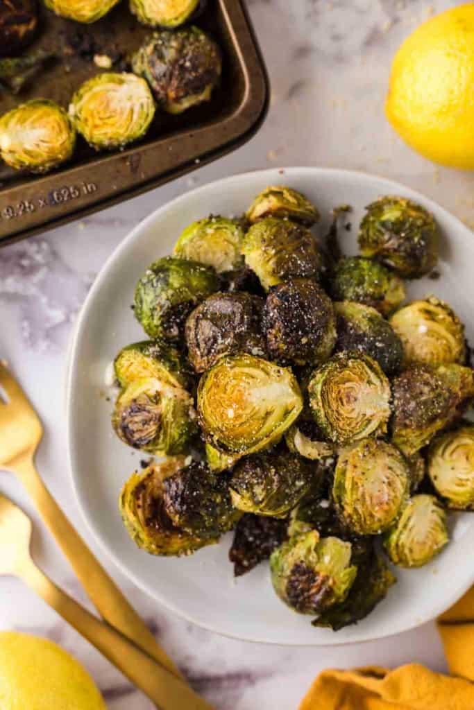 Oven Baked Brussels Sprouts on a round white plate next to whole lemons and golden forks.