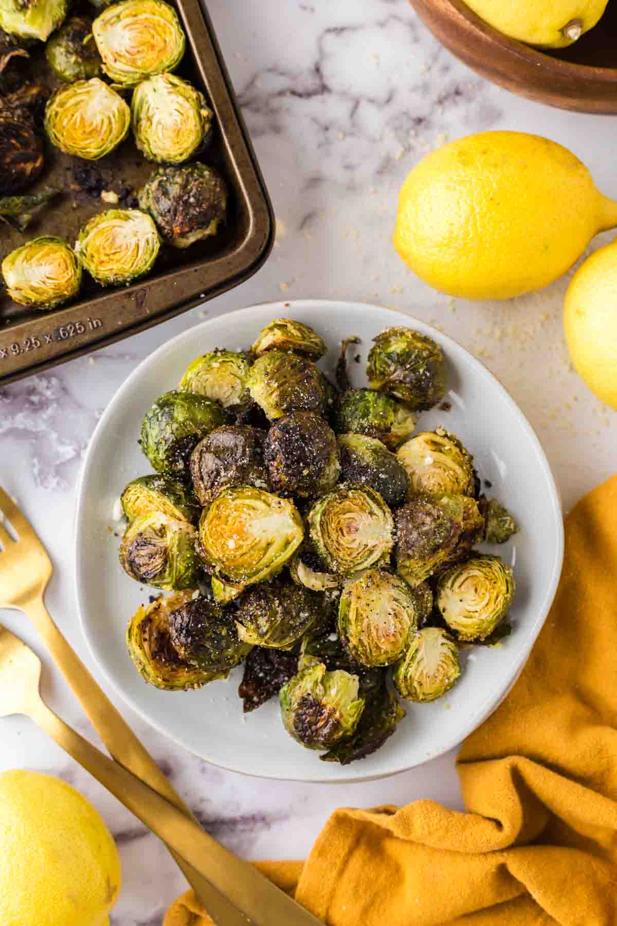 Oven Baked Brussels Sprouts on a round white plate next to whole lemons and golden forks.