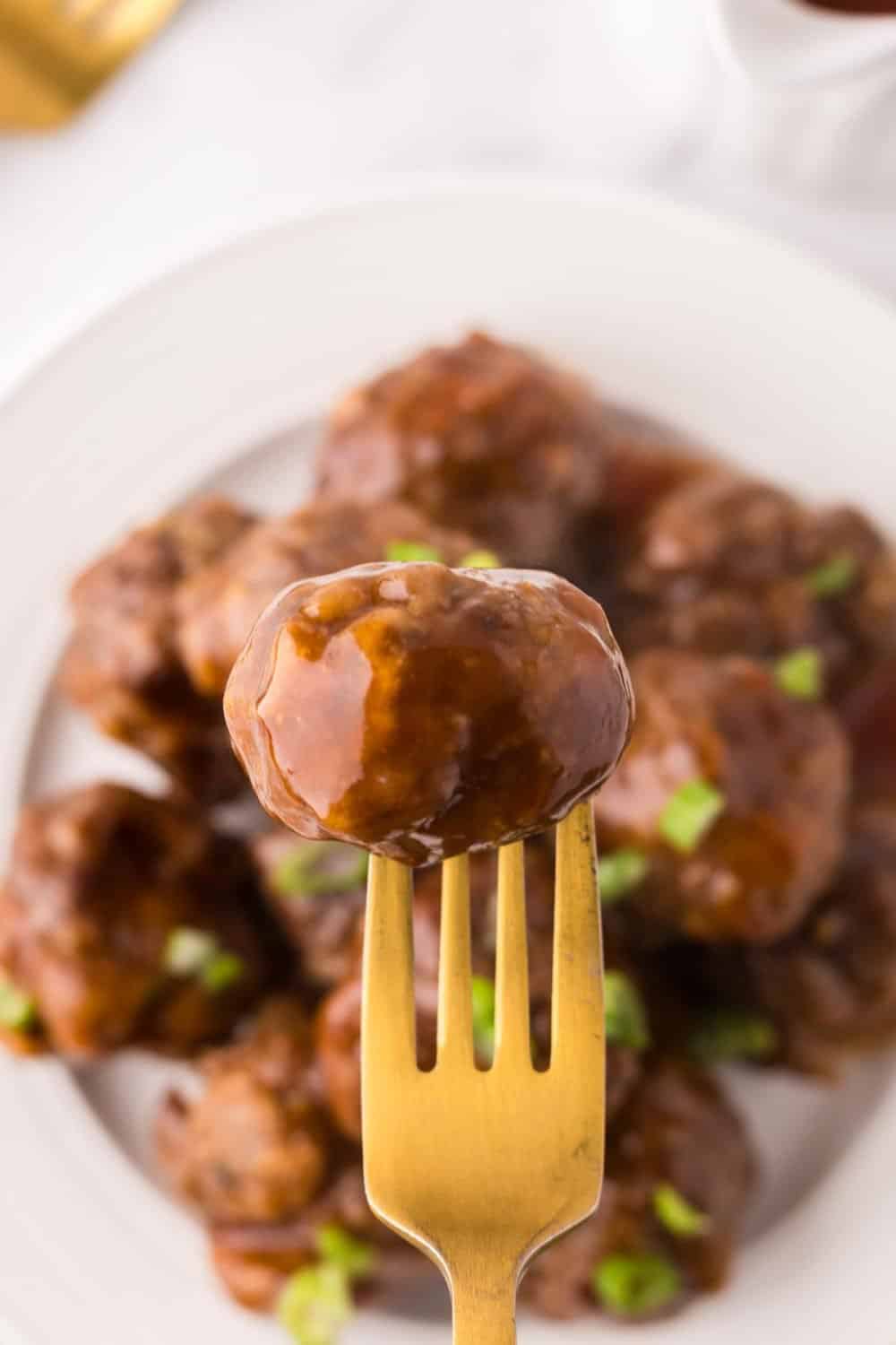 Golden fork stabbed into one sweet and sour meatball over a stack on a round white plate.