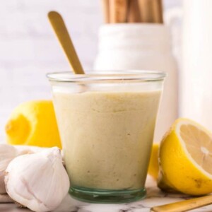 tahini dressing in a small glass jar with lemons and garlic all around with a gold spoon in it.