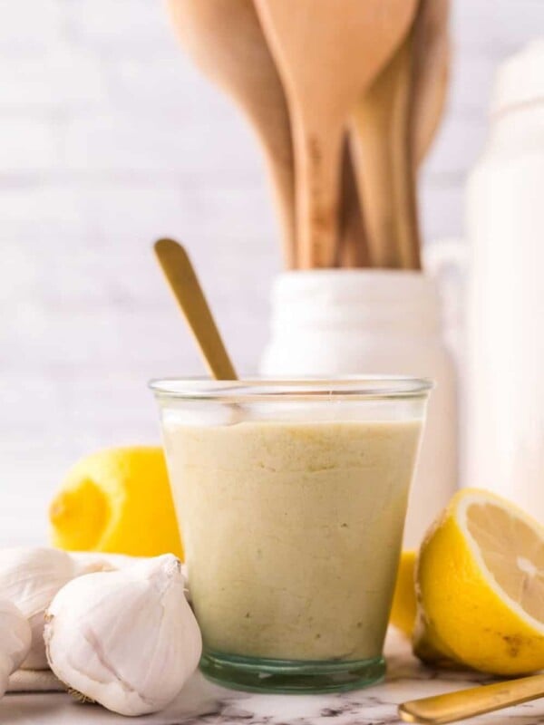 tahini dressing in a small glass jar with lemons and garlic all around with a gold spoon in it.