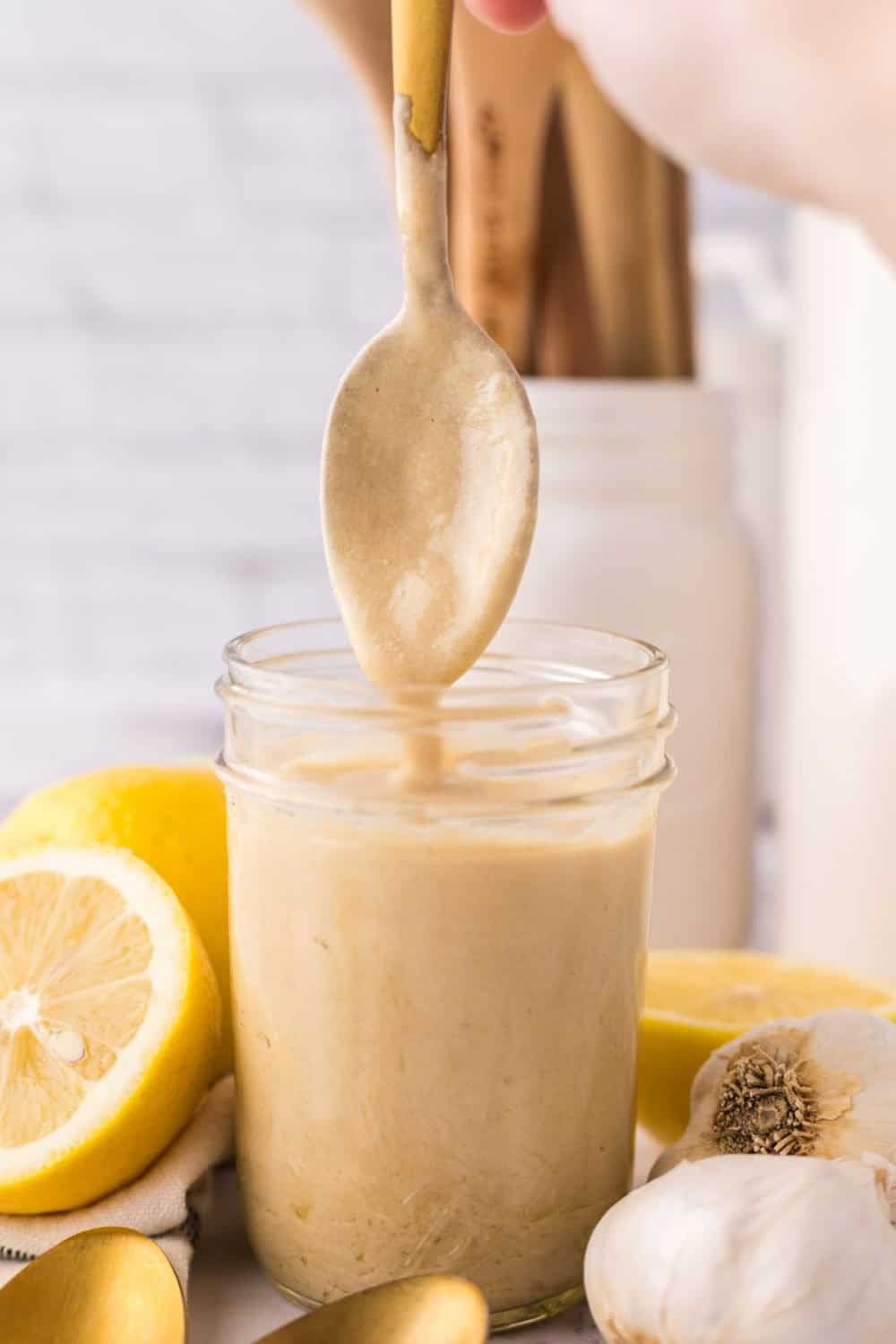 tahini dressing dripping off a spoon over a small glass mason jar with lemons and garlic all around.