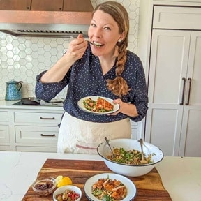 A smiling woman in a kitchen eats from a small plate, holding a fork. She stands behind a counter with bowls of food, a lemon, and olives, wearing a dark blouse and a light apron.