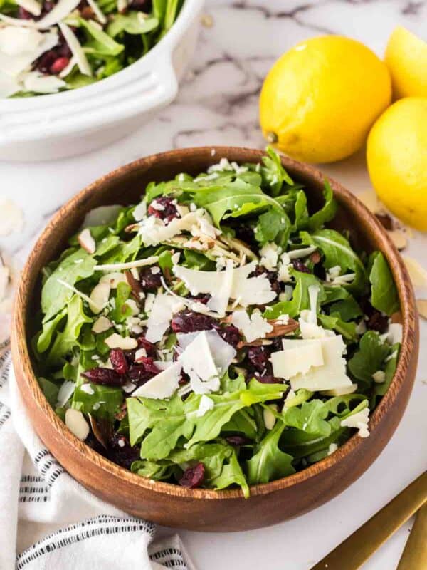 arugula salad recipe in a round wooden serving bowl with golden forks and lemons to the side.
