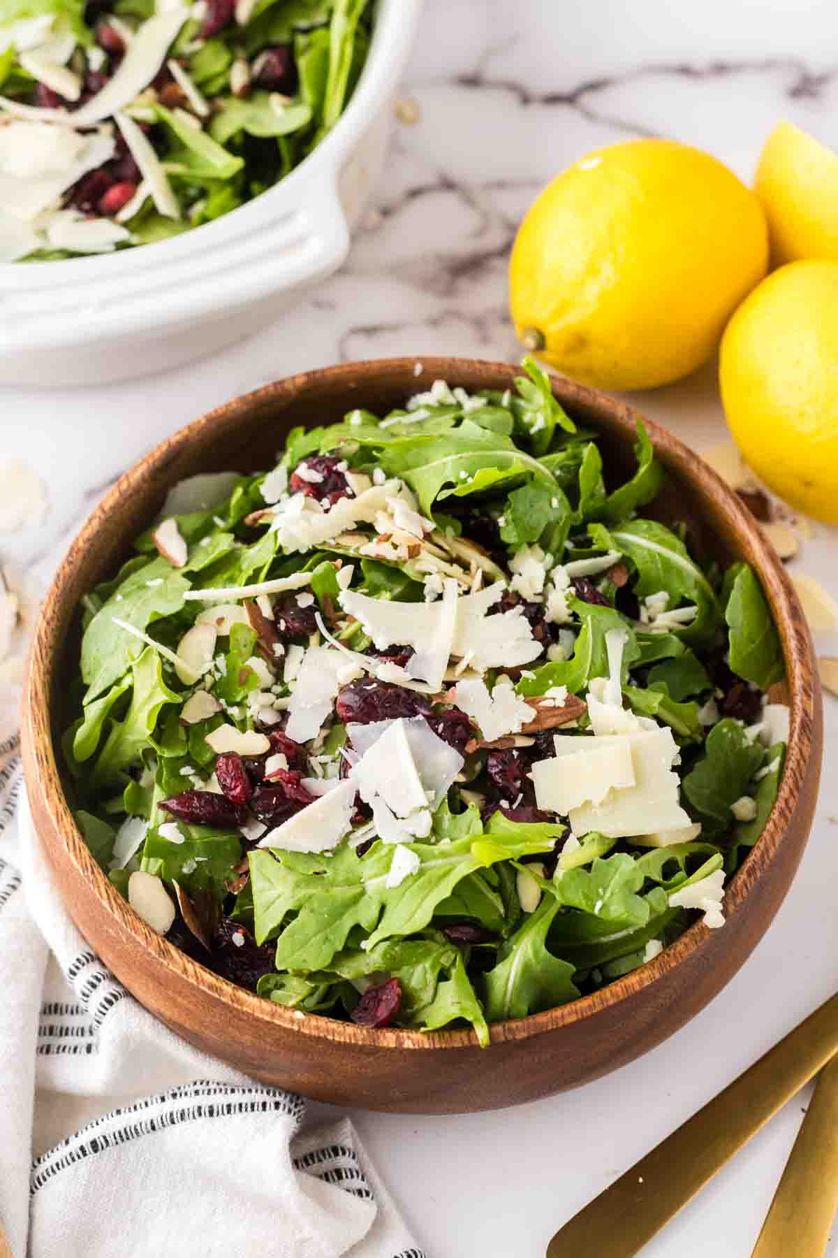 arugula salad recipe in a round wooden serving bowl with golden forks and lemons to the side.