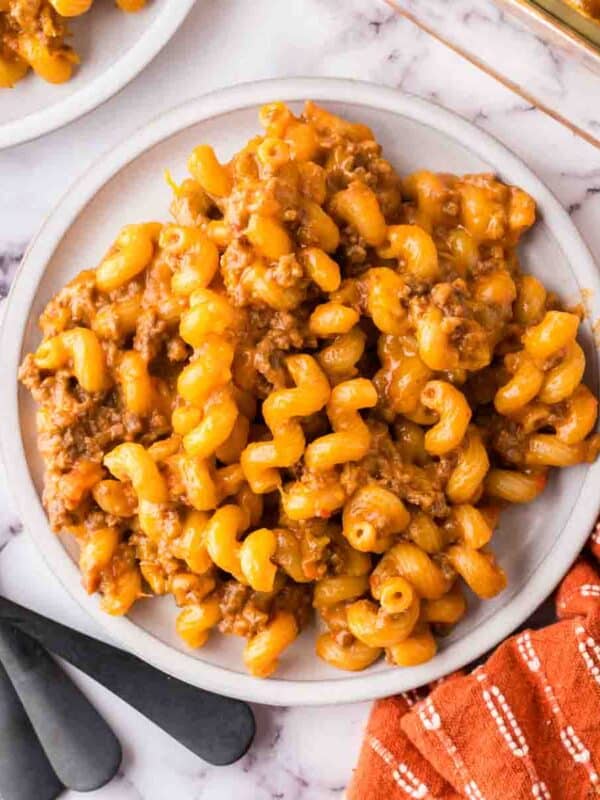 A plate of cheesy macaroni with ground beef, reminiscent of cheeseburger casserole, sits on a marble surface next to black forks and an orange checkered napkin. A casserole dish with more pasta is partially visible in the corner.