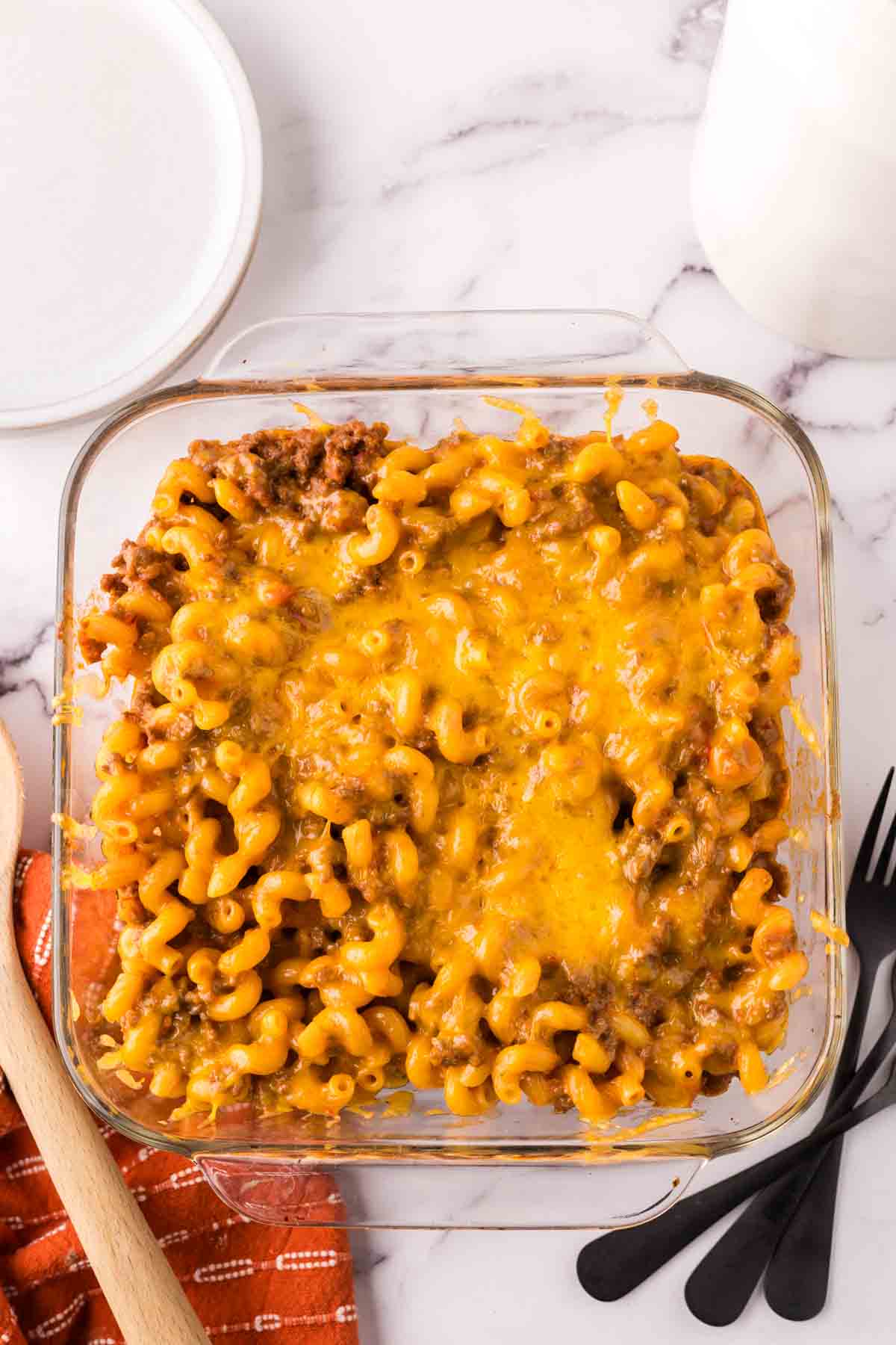 A glass baking dish filled with cheeseburger casserole&mdash;baked macaroni and cheese topped with melted cheddar and ground beef&mdash;sits next to a wooden spoon, black utensils, a red napkin, a white plate, and a glass of milk on a marble surface.
