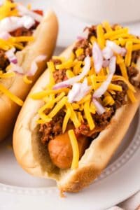 chili dogs recipe over round white plates with cheese and onions on top.