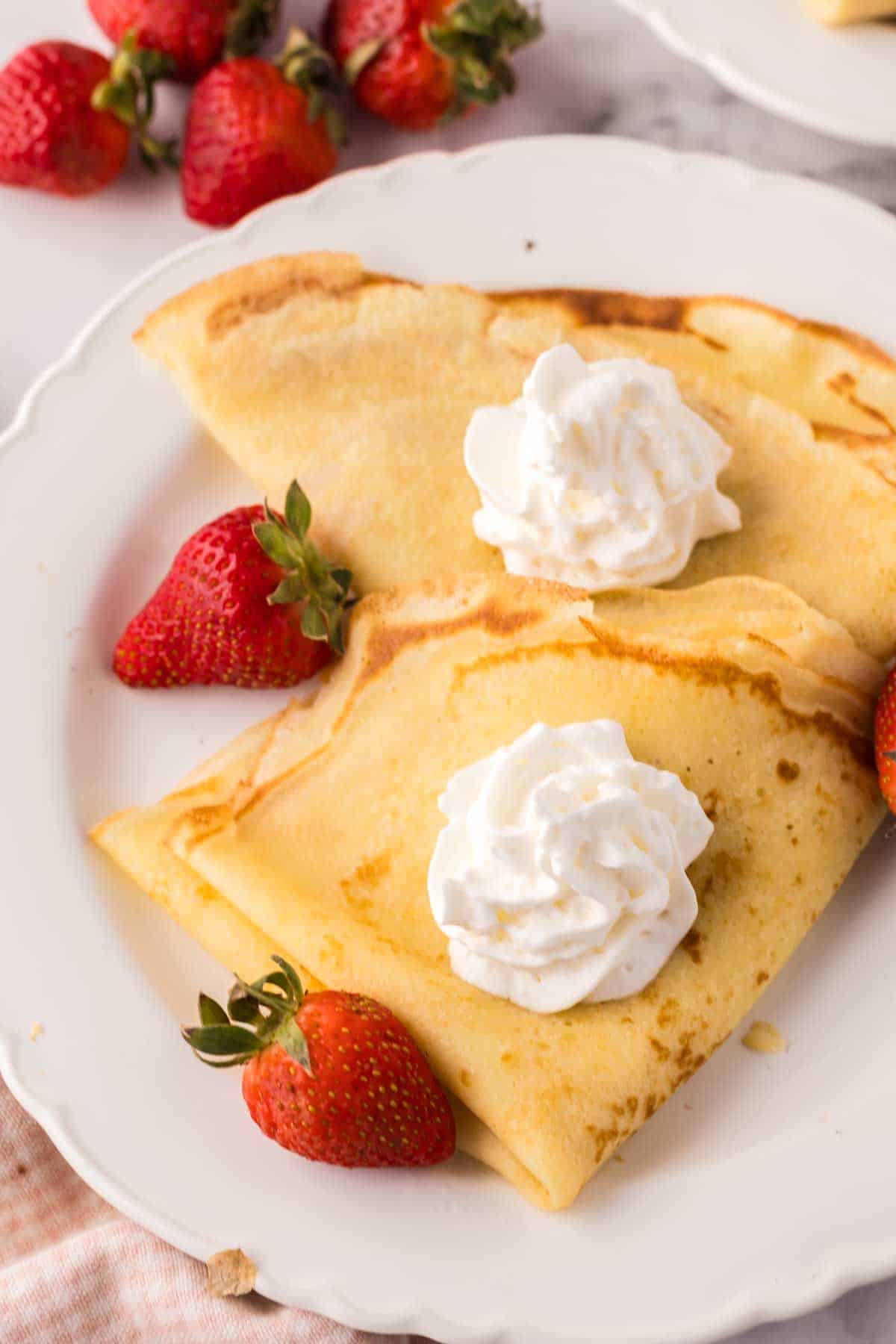 folded crepes on a white plate with fresh berries around and whipped cream.
