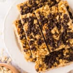 A plate stacked with several healthy homemade granola bars topped with chocolate chips. Oats and pretzel pieces are visible in the bars, with a bowl of oats and a striped cloth nearby.