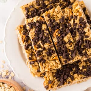 A plate stacked with several healthy homemade granola bars topped with chocolate chips. Oats and pretzel pieces are visible in the bars, with a bowl of oats and a striped cloth nearby.