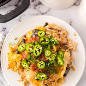 a round white plate with skillet, shredded chicken nachos with melted cheese and jalapeños on top.