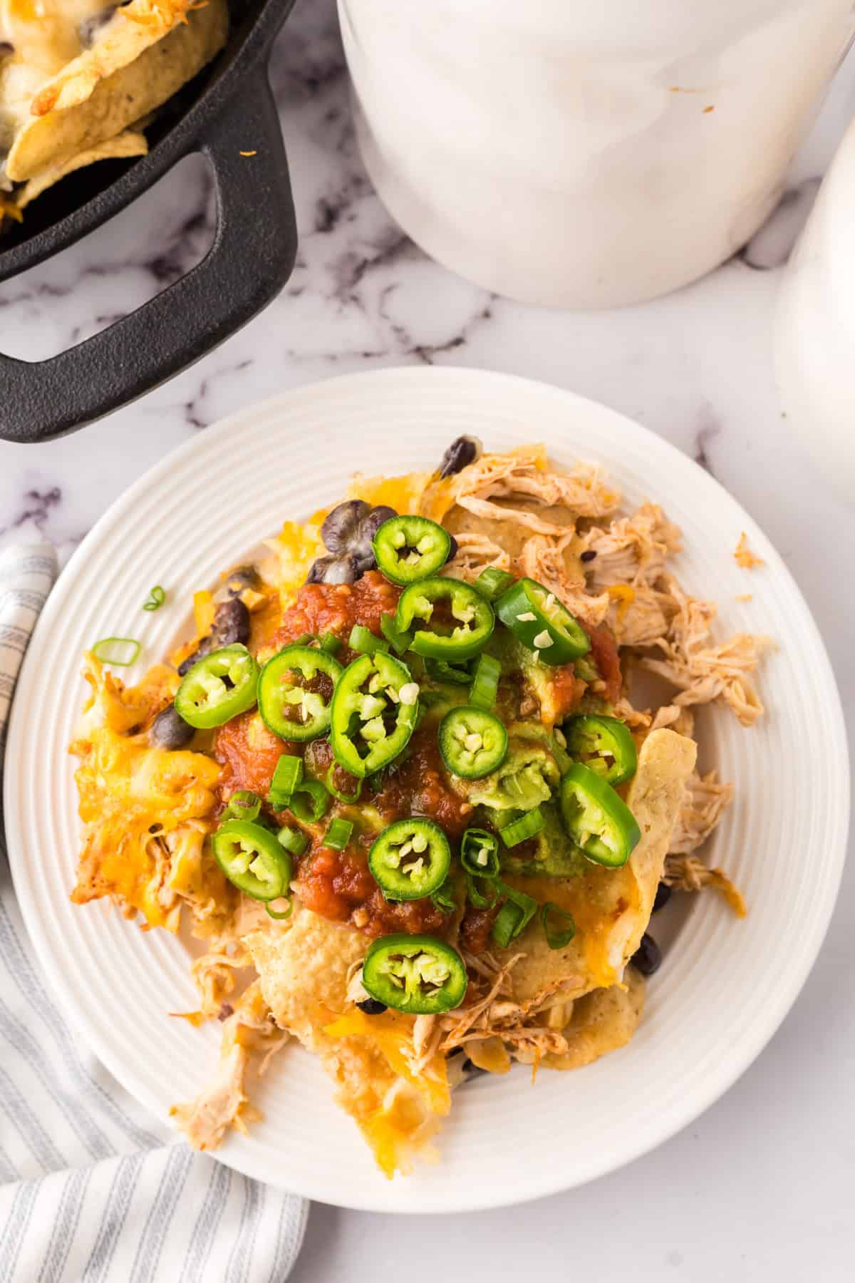 a round white plate with skillet, shredded chicken nachos with melted cheese and jalapeños on top.