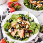 spinach and berry salad on a round white plate with black forks to the side.