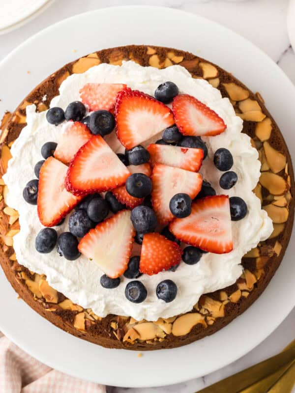 A round honey cake recipe is topped with whipped cream, sliced strawberries, blueberries, and almond slices on a white plate. Nearby are three gold forks, a white spatula, and a pink checkered napkin.