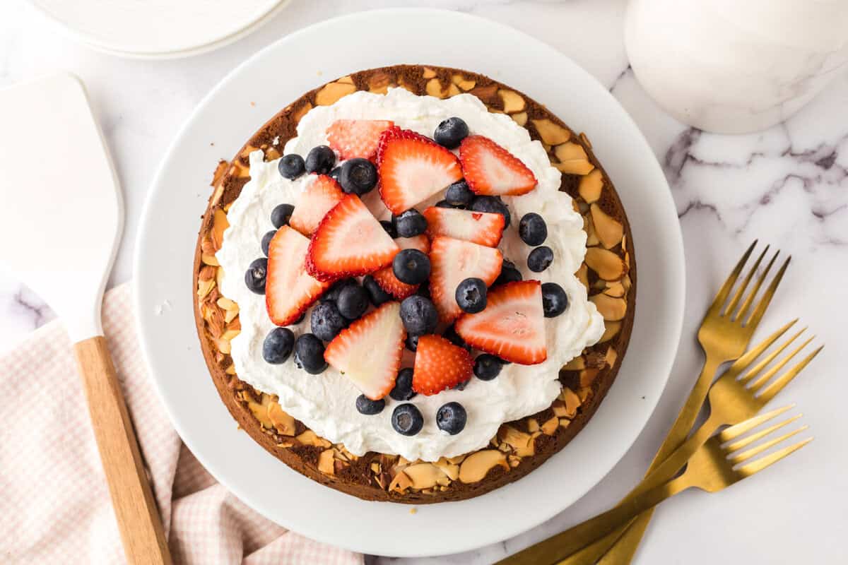 A round honey cake recipe is topped with whipped cream, sliced strawberries, blueberries, and almond slices on a white plate. Nearby are three gold forks, a white spatula, and a pink checkered napkin.