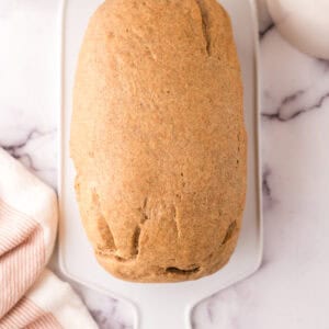 A loaf from a whole wheat bread recipe sits on a white rectangular cutting board, surrounded by a pink-striped towel and white containers on a marble surface.