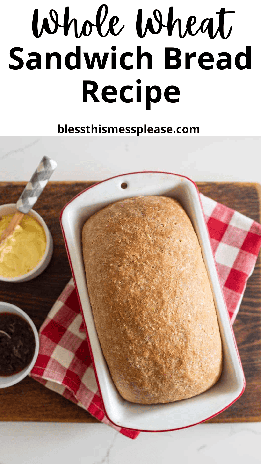 A loaf of whole wheat sandwich bread recipe sits in a white baking dish on a red and white checkered cloth, accompanied by a bowl of butter and a bowl of jam on a wooden board.