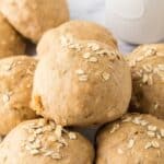 A close-up of freshly baked whole wheat hamburger buns topped with oats. The buns are stacked on a marble surface with a hint of a white, round container in the background.