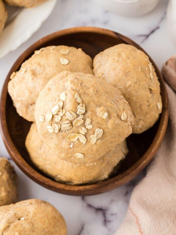 A wooden bowl filled with freshly baked, round whole wheat hamburger buns topped with oats. The buns are placed on a marble surface, with more rolls visible nearby and a beige cloth partially in view.