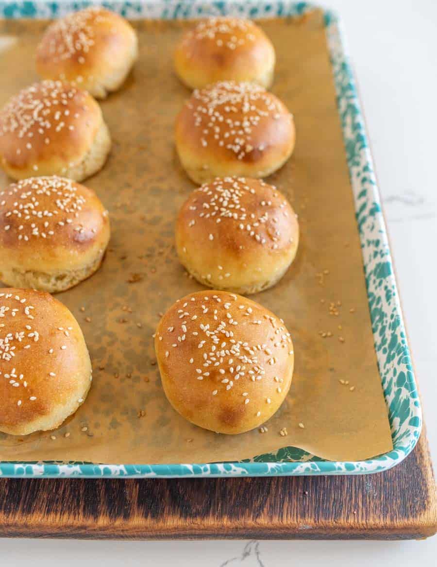 A baking tray lined with parchment paper holds eight golden-brown, round brioche buns topped with sesame seeds. The tray sits on a wooden board.