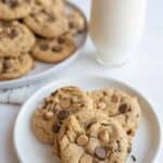 A small plate with three peanut butter chocolate chip cookies sits in the foreground, accompanied by a glass of milk. In the background, more cookies are piled on a larger plate. The scene is set on a light-colored surface.
