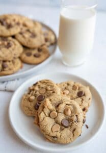 A small plate with three peanut butter chocolate chip cookies sits in the foreground, accompanied by a glass of milk. In the background, more cookies are piled on a larger plate. The scene is set on a light-colored surface.
